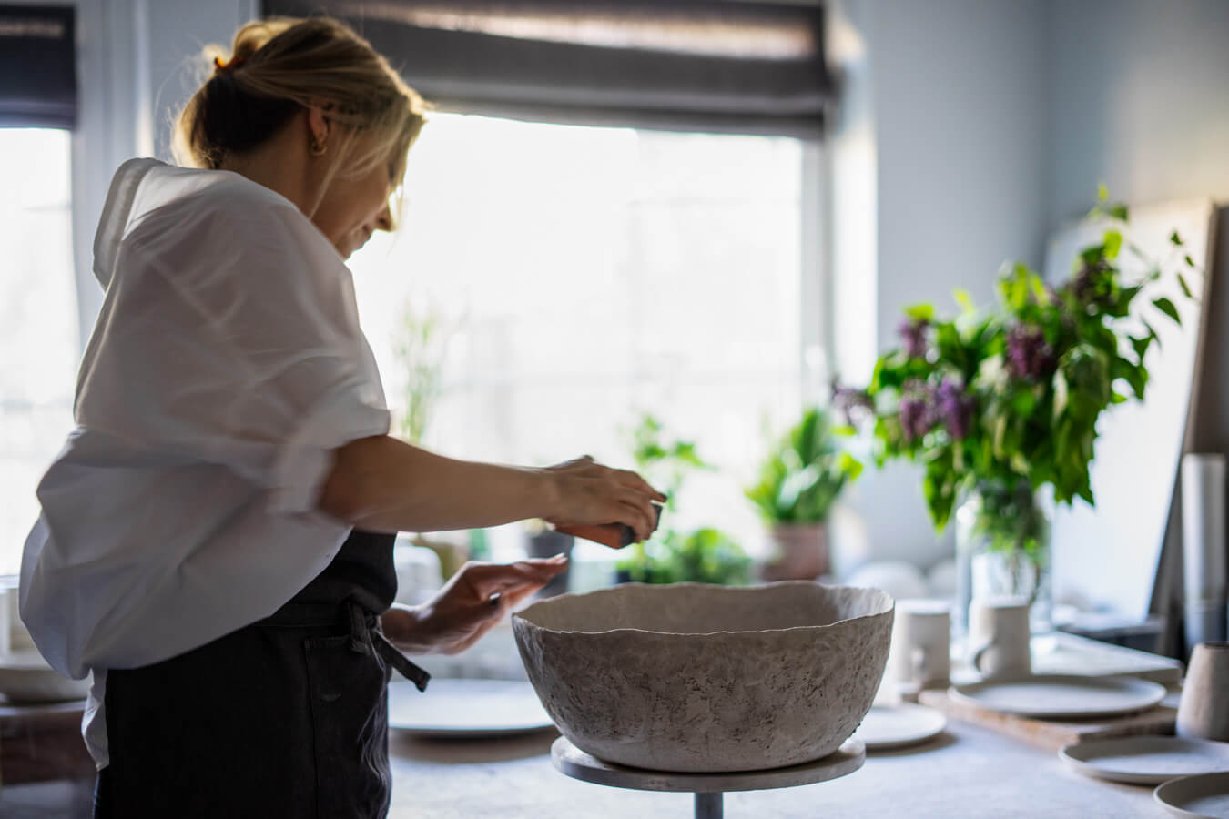 Kasia Białek przy pracy w pracowni — wygładza dużą fakturowaną misę, w oknie bzy